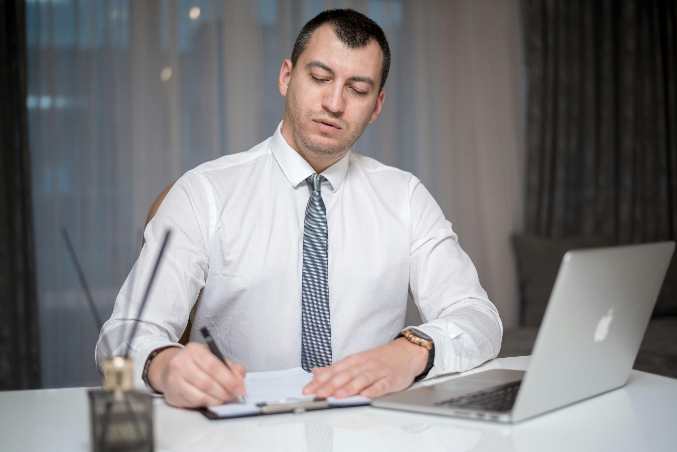 professionally dressed man writing at a desk with an open laptop